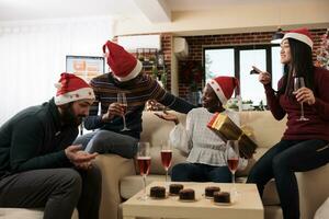 glad upphetsad afrikansk amerikan kvinna anställd innehav xmas gåva och gissa hemlighet santa i kontor. olika kollegor dricka alkohol och delning jul presenterar foto