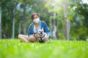 en kvinna som bär en medicinsk mask sitter på gräsmattan med en vit fransk bulldog. foto