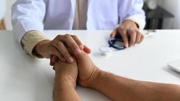 en läkare i en labbdräkt med ett stetoskop håller patientens hand för att trösta, doktorn tar tag i patientens hand för att uppmuntra. foto