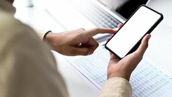 ung man som använder smartphone i handen med dokument och bärbar dator på sitt skrivbord. foto