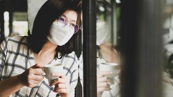 en kvinna i en medicinsk mask som håller en kaffekopp tittar ut genom fönstret.