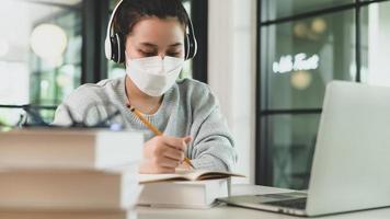 asiatisk tjej med stetoskop bär medicinsk mask som tar anteckningar och studerar online med bärbar dator.