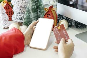 mild händer vagga en smartphone med en vit skärm falsk och håll en kreditera kort till affär uppkopplad upp förbi de festlig scen av en skön jul träd Utsmyckad, med jul bollar, och tall kottar. foto