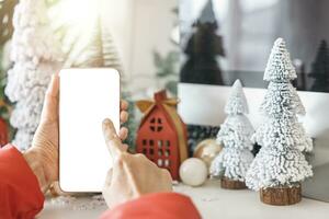 mild händer vagga en smartphone med en vit skärm falsk och fingrar på de skärm till affär uppkopplad upp förbi de festlig scen av en skön jul träd Utsmyckad, med jul bollar, och tall kottar. foto