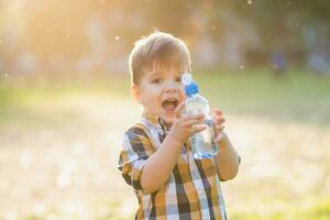 glad barn drycker klar vatten från en flaska på en solig dag i natur foto