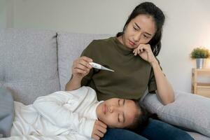 asiatisk kvinna ta vård av liten sjuk dotter. sjuk barn liggande på säng under filt, med orolig. enda mamma tar vård av sjuk dotter på Hem. barn har en hög feber. omslag på de soffa och sjuk foto