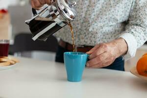 senior man använder sig av franska Tryck och häller varm dryck efter framställning kaffe i kök för frukost. äldre person i de morgon- njuter färsk brun Kafé espresso kopp koffein från årgång råna foto