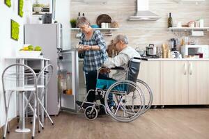 senior kvinna framställning frukost för handikappade Make tar ägg kartong från kylskåp , levande med man med gående funktionshinder. Inaktiverad senior manlig i rullstolshjälp hans fru i kök. foto