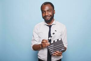 afrikansk amerikan film direktör innehav öppen clapperboard i händer ser självsäker medan Framställ för kamera. ung framgångsrik filmskapare gripande kläpp stående stolt mot blå bakgrund. foto