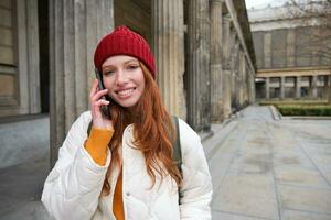 leende rödhårig kvinna turist samtal på mobil telefon och promenader runt om stad. Lycklig studerande i röd hatt samtal vän, står på gata och användningar smartphone foto
