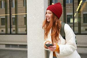 eleganta urban flicka använder sig av mobil telefon app, stående i stad, väntar för taxi, ser på smartphone Ansökan, textning meddelande foto