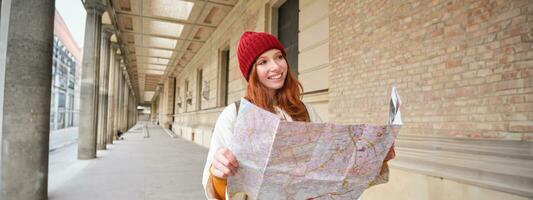 leende ung rödhårig kvinna i röd hatt, utseende på papper Karta till se för turist attraktion. turism och människor begrepp. flicka utforskar stad, försökte till hitta sätt foto