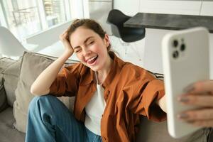 porträtt av skön, Lycklig ung kvinna modell, Sammanträde på soffa med smartphone, tar selfie på mobil telefon, framställning glad leende ansikte foto