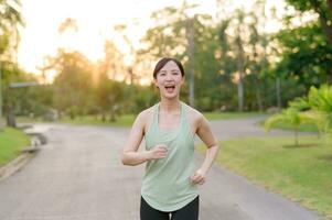 passa asiatisk ung kvinna joggning i parkera leende Lycklig löpning och njuter en friska utomhus- livsstil. kvinna joggare. kondition löpare flicka i offentlig parkera. friska livsstil och wellness varelse begrepp foto