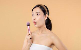 porträtt Lycklig ung asiatisk kvinna med naturlig göra upp ansikte innehav kosmetisk hud pulver rodnad isolerat på beige bakgrund. kvinna tillämpa hudvård borsta behandling. skönhet produkt kosmetologi begrepp. foto