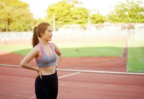 ung asiatisk kvinna avkopplande efter träna och joggning på de fotboll stadion i de morgon- med Sol blossa bakgrund. foto