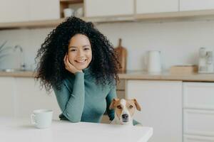 glad kvinna med lockigt hår och henne hund på en kök tabell, delning en ögonblick av lycka foto
