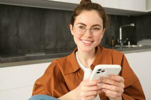 stänga upp porträtt av leende ung kvinna, kvinna modell Sammanträde i kök med smartphone, använder sig av mobil telefon och leende foto