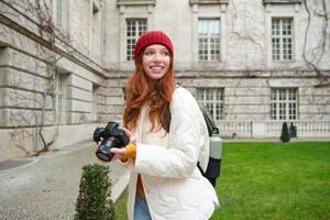 Lycklig rödhårig flicka turist, tar foton, fotograf med professionell kamera promenader runt om stad och fångar skön bilder foto