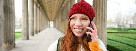 porträtt av rödhårig europeisk flicka i röd hatt, gör en telefon ringa upp, promenader i stad och samtal till vän på smartphone foto