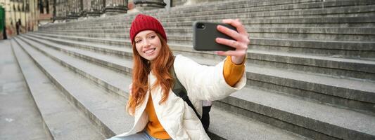 ung rödhårig turist tar selfie i främre av museum på trappa, innehar smartphone och utseende på mobil kamera, gör Foto av själv med telefon