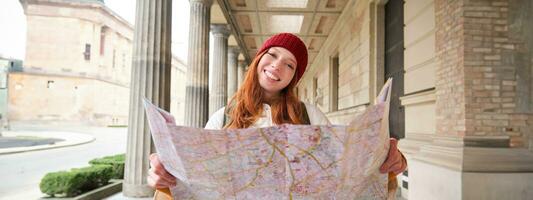 äventyrlig rödhårig flicka promenader i stad med papper Karta, utforskar stad som turist, utseende för populär turism attraktioner, utseende runt om upphetsad och ler foto