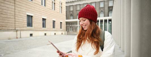 eleganta ingefära flicka, turist promenader med digital läsplatta runt om stad, kvinna ansluter till iternet på henne grej, ser upp information, textning meddelande foto