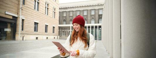 eleganta ingefära flicka, turist promenader med digital läsplatta runt om stad, kvinna ansluter till iternet på henne grej, ser upp information, textning meddelande foto