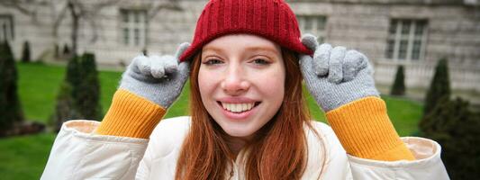 söt flicka studerande i röd hatt, värma handskar, sitter i parkera, ler och utseende Lycklig. foto