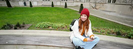 ung eleganta kvinna studerande med ryggsäck, sitter på bänk och användningar smartphone, spelar på mobil telefon foto