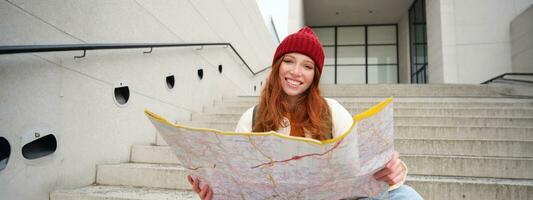 ung leende rödhårig flicka, turist sitter på trappa utomhus med stad papper Karta, ser för riktning, resande backpacker utforskar stad och utseende för sightseeing foto