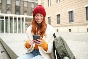 ung rödhårig kvinna med smartphone, Sammanträde utomhus med ryggsäck, studerande ser på henne mobil telefon, texter meddelande foto