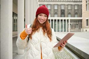 eleganta ingefära flicka, turist promenader med digital läsplatta runt om stad, kvinna ansluter till iternet på henne grej, ser upp information, textning meddelande foto