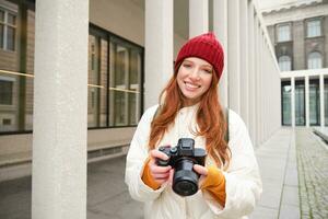 leende rödhårig flicka fotograf, tar bilder i stad, gör foton utomhus på professionell kamera