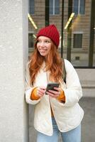 eleganta urban flicka använder sig av mobil telefon app, stående i stad, väntar för taxi, ser på smartphone Ansökan, textning meddelande foto