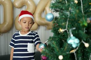 glad jul 2024 begrepp asiatisk pojke dekorerar jul träd med färgrik jul bollar. foto