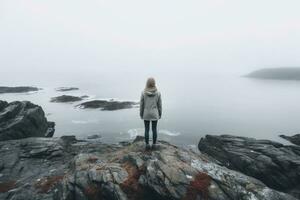 ai genererad ung kvinna stående på en sten ser på de hav i de dimma, kvinna ensam ser på dimmig hav reser äventyr livsstil utomhus- ensamhet ledsen känslor vinter- ner, ai genererad foto