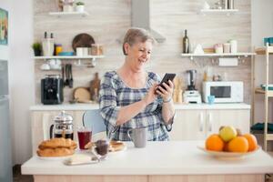 gammal kvinna bläddring på internet använder sig av smartphone i kök under frukost. . uppkopplad kommunikation ansluten till de värld, senior fritid tid med grej på pensionering ålder foto