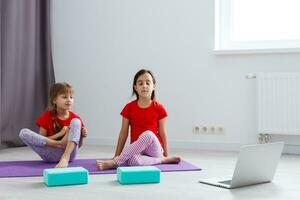 två liten flickor praktiserande yoga, stretching, kondition förbi video på anteckningsbok. avlägsen uppkopplad utbildning Träning, aerob på Hem. friska livsstil, coronavirus, stanna kvar Hem. barn sport Hem karantän. foto