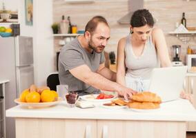 Make och fru läsning en post på bärbar dator i de morgon- under frukost. gift Make och fru i pyjamas använder sig av internet webb uppkopplad modern teknologi, leende och Lycklig i de morgon. läsning Nyheter bläddring internet foto