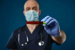 läkare i mask, blå handskar, medicinsk skrubba. innehav röd laboratorium testa rör med grön keps i hand. blå bakgrund. coronavirus, covid19. stänga upp foto