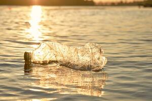 skrynkliga plast flaska flyter på de yta av de vatten. solnedgång, grön träd. människor och ekologi. flodstrand förorening. närbild skott. foto