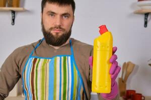 ung skäggig man i kök gör reklam. han främjar rengöringsmedel, innehav den i ett hand och visar på kamera foto