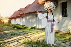 brunett flicka i en vit ukrainska äkta nationell kostym och en krans av blommor är Framställ mot en vit hydda. foto