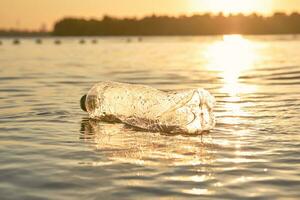 skrynkliga plast flaska flyter på de yta av de vatten. solnedgång, grön träd. människor och ekologi. flodstrand förorening. närbild skott. foto