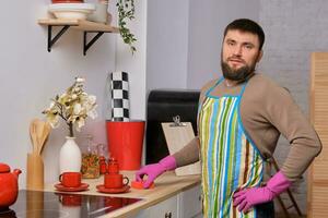 ung stilig skäggig man i de kök, bär förkläde och rosa handskar rengör de matlagning yta använder sig av tvättmedel foto