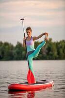 sportig kvinna i yoga placera på paddelbräda, håller på med yoga på supera styrelse, övning för flexibilitet och stretching av muskler foto