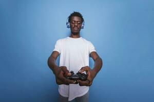 afrikansk amerikan video gamer i studio använder sig av kontrollant och hörlurar. svart man spelar ett internet spel på en trösta med en joystick medan stående över isolerat blå bakgrund. foto