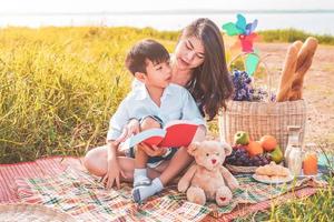 vacker asiatisk mor och son som gör picknick och läser berättelse i röda böcker i påsksommarfesten på äng nära sjö och berg. semester. människors livsstil och lyckliga familjeliv. thailändsk person