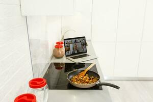 skön flicka är inlärning till laga mat friska mat uppkopplad förbi de internet från en bärbar dator i grå kök på tabell. foto